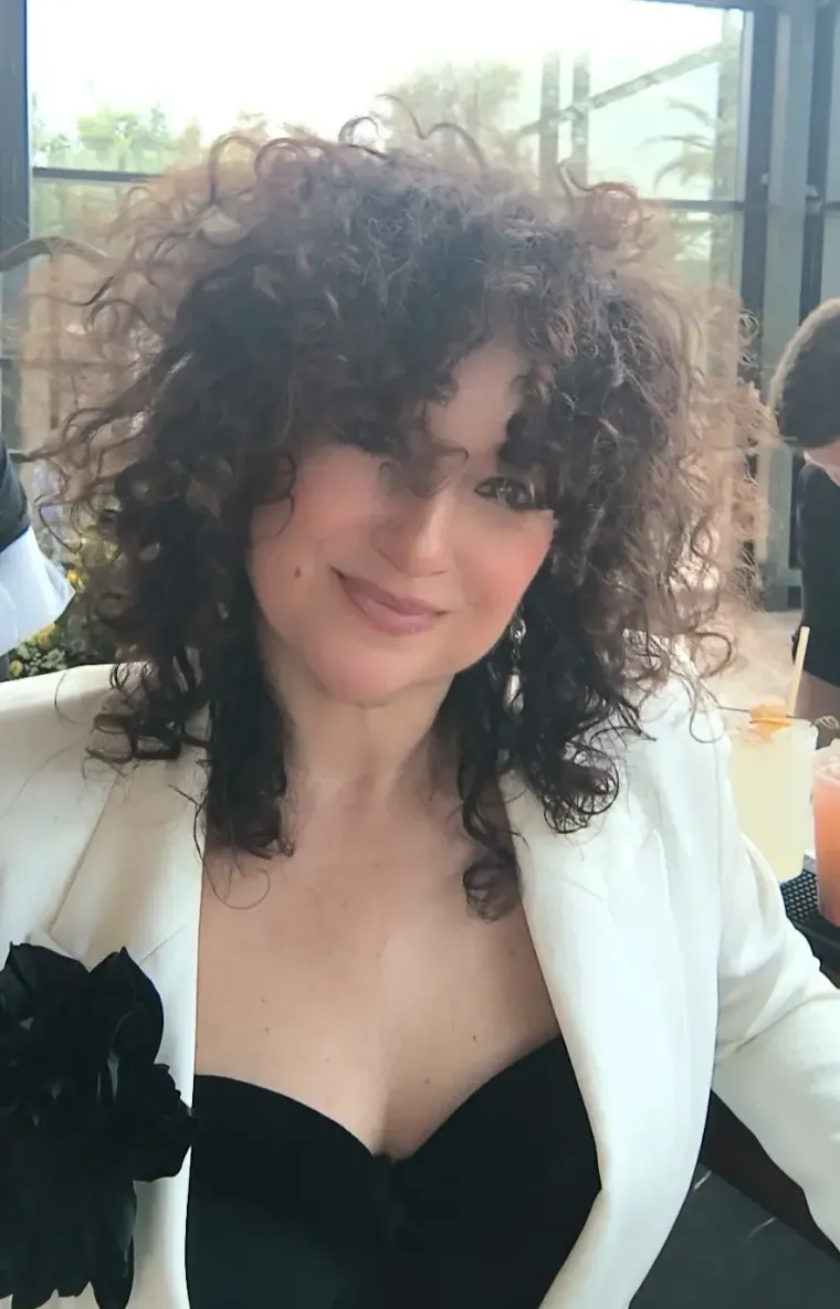 Mujer con cabello castaño rizado, vestida con un blazer blanco, sonriendo.