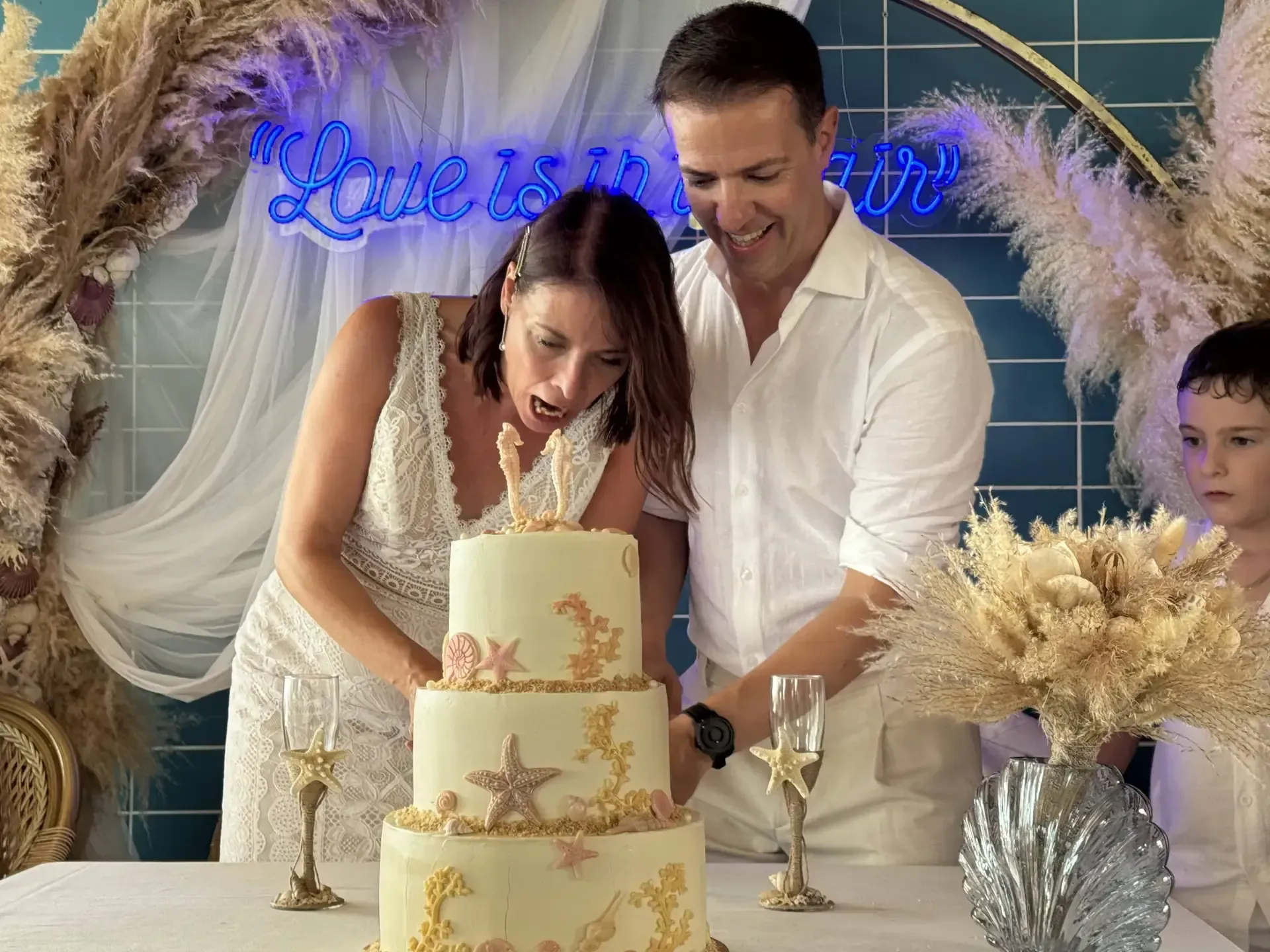 Pareja cortando un pastel de bodas con temática oceánica; letrero de neón 