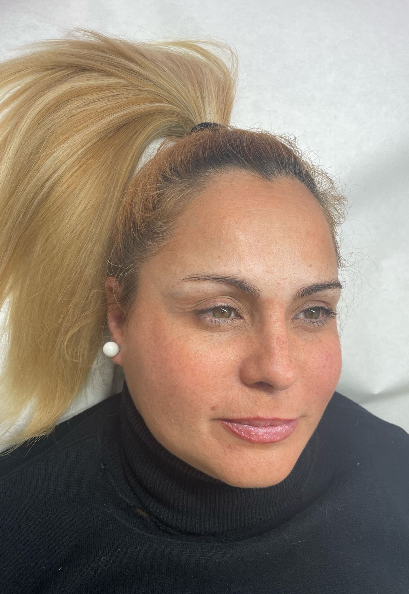 Una mujer con cabello rubio recogido en una cola de caballo lleva un cuello alto negro y pendientes.