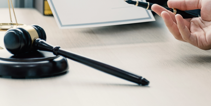 Gros plan sur un maillet de juge posé sur sa base, sur une surface en bois. En arrière-plan, une main tient un stylo au-dessus d’un document, suggérant un contexte juridique ou administratif.