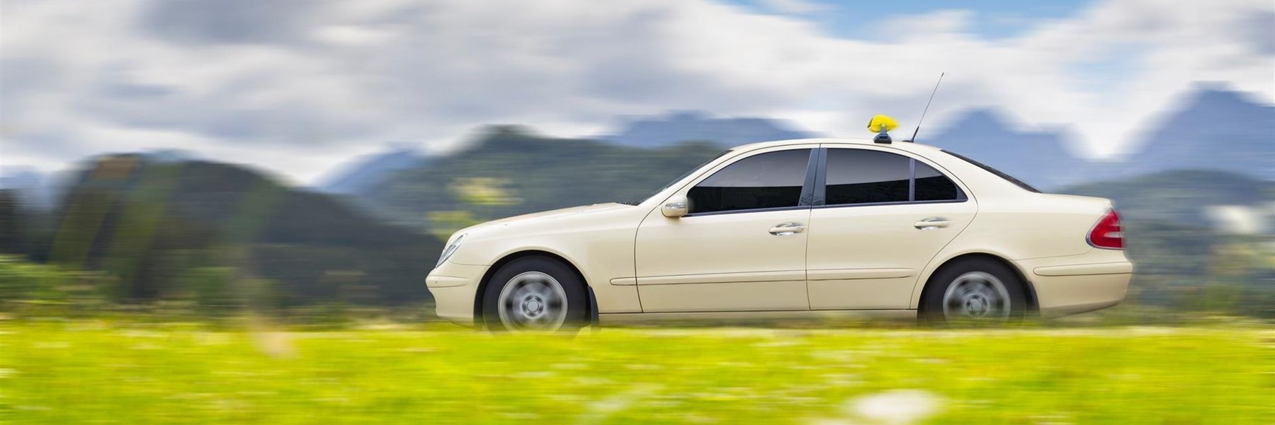 Ein weißes Auto fährt eine Straße entlang, im Hintergrund sind Berge zu sehen.