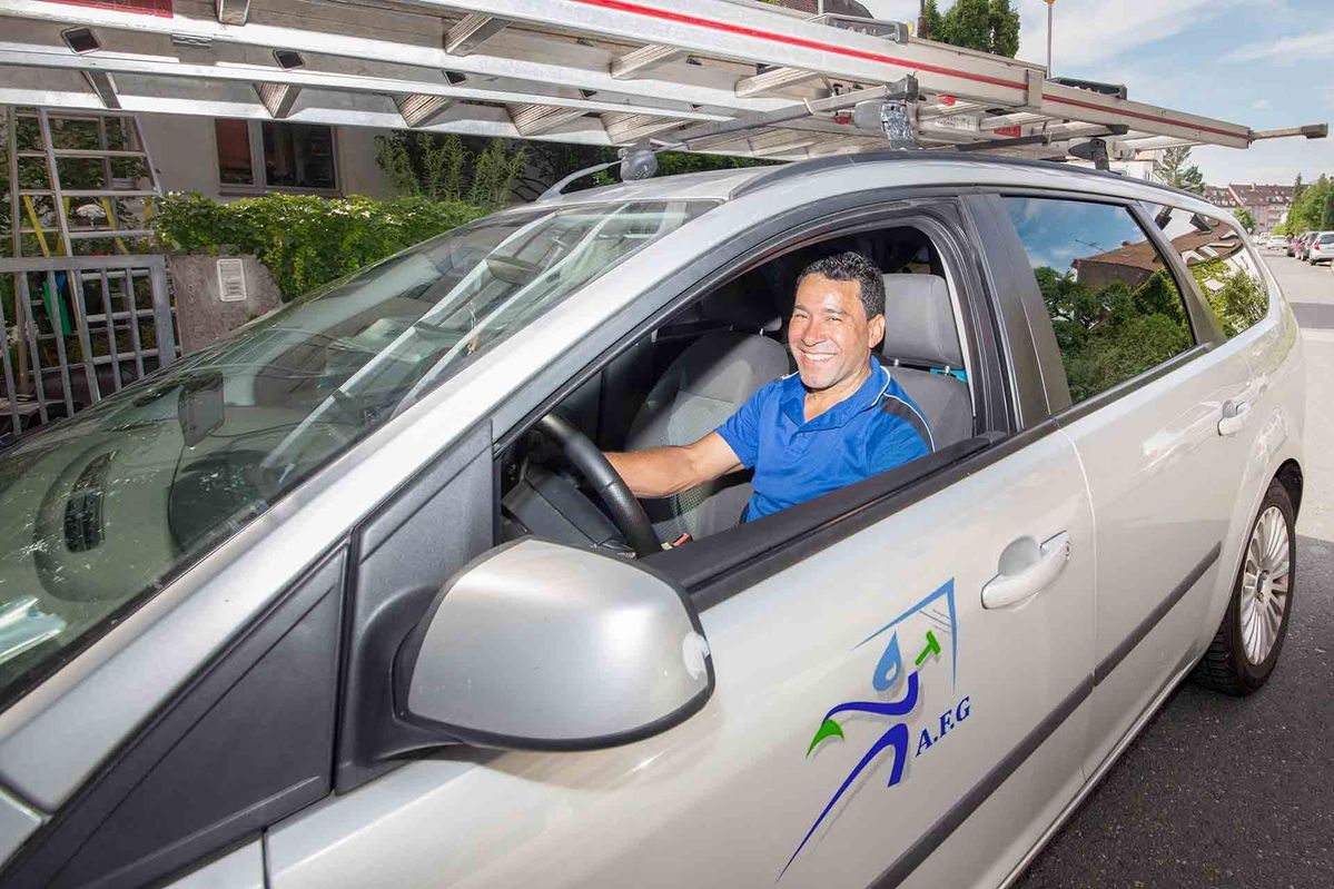 Mann in einem silbernen Auto mit einer Leiter auf dem Dach, lächelnd. Logo an der Seite.