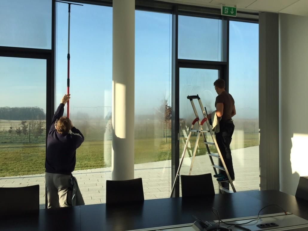 Zwei Personen putzen große Fenster in einem Büro mit malerischer Aussicht. Einer benutzt eine Stange, der andere eine Leiter.
