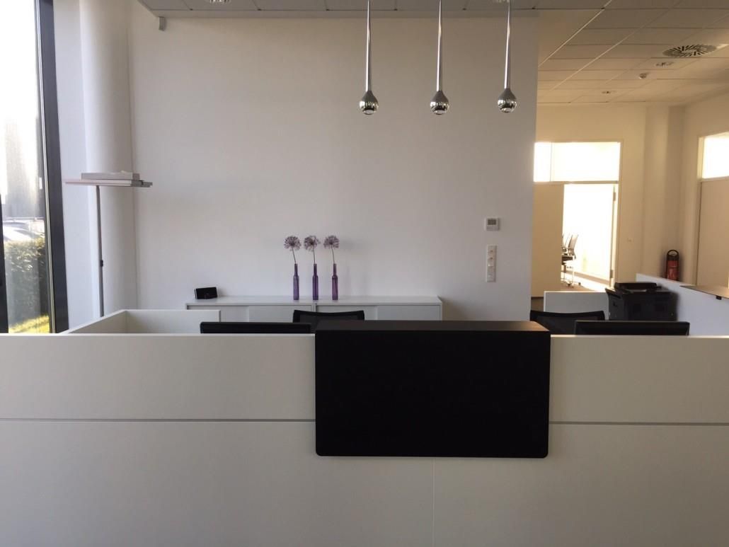Weiß-schwarzer Empfangstresen in einem hellen Büroraum. Drei Pendelleuchten hängen darüber.