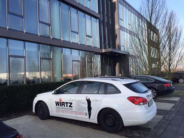 Weißes Auto mit „Wirtz“-Logo parkt vor einem modernen Bürogebäude mit reflektierenden Fenstern.