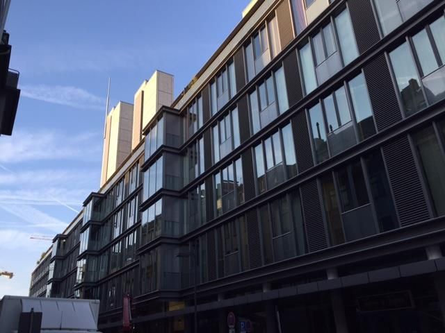 Modernes Gebäude mit mehreren Fenstern, grauer Fassade und einem Turm im Hintergrund. Sonniger Tag.