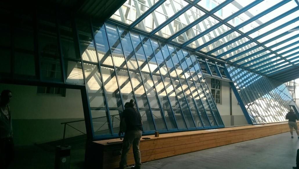 Großes Gebäudeinneres mit Glaswänden und Oberlicht, in dem Menschen arbeiten. Blaue Akzente, Holzvertäfelung entlang des Sockels.