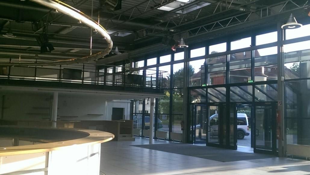 Innenraum eines großen, leeren Veranstaltungsortes mit Bar, Balkon und Glaswänden. Sonnenlicht fällt herein.