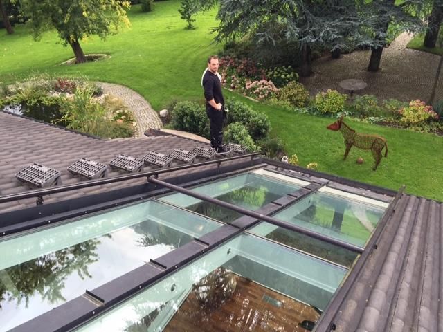 Mann steht auf einem Dach mit Glasplatten und blickt auf einen grünen Rasen mit einem digital hinzugefügten braunen Hund.
