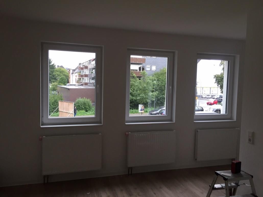Drei Fenster in einem Raum, darunter jeweils ein Heizkörper. Draußen gibt es einen Blick auf Gebäude und Bäume.