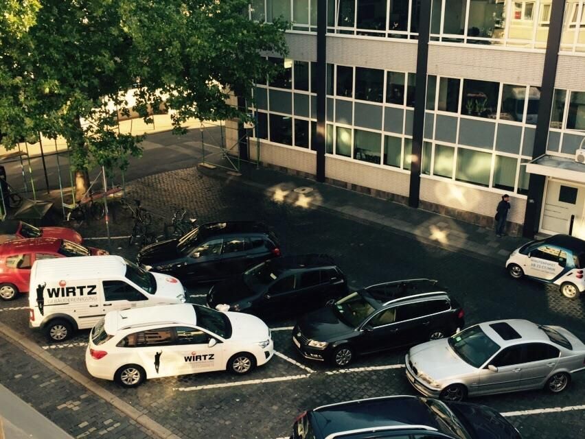 Vor einem Gebäude mit Fenstern geparkte Autos; weißer Wirtz-Lieferwagen und Autos; links ein Baum.