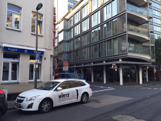 Straßenszene mit einem geparkten weißen Kombi, rechts ein mehrstöckiges Gebäude und links ein kleines Gebäude.