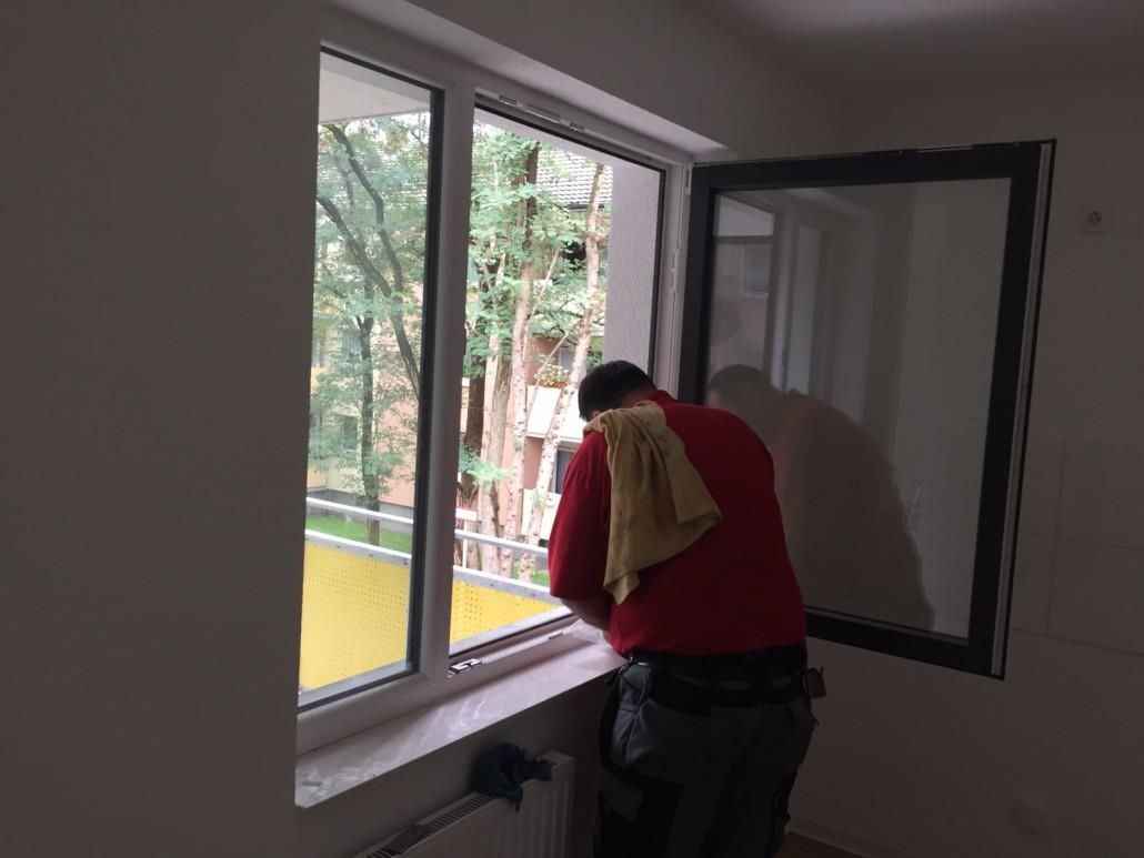 Person putzt Fenster, Innenaufnahme. Er trägt Rot, draußen sind Bäume zu sehen.