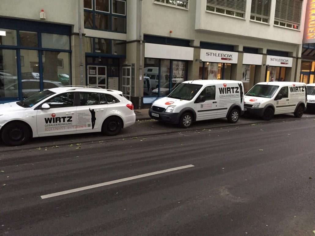 Weiße Autos und Lieferwagen mit der Aufschrift „WIRTZ“ parken auf einer Straße vor Ladenfronten.
