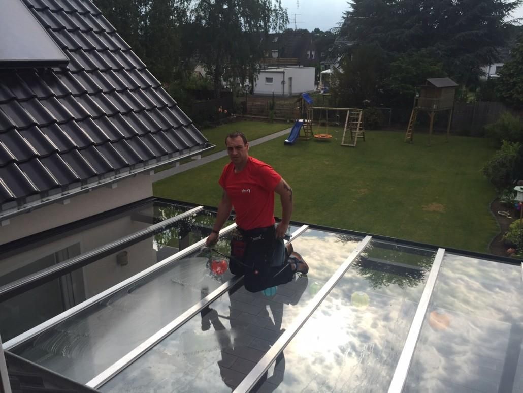 Mann im roten Hemd reinigt Glasdach; im Hintergrund Haus und grüner Garten.