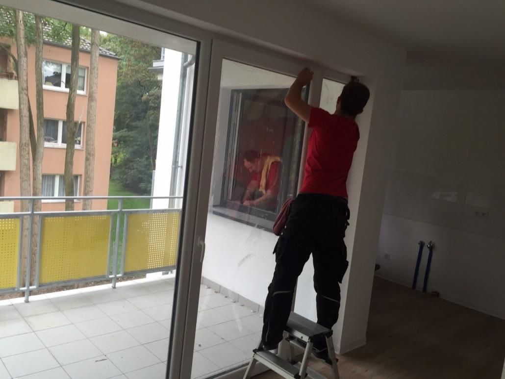 Mann auf einer Trittleiter installiert etwas über einer Glasschiebetür. Balkon mit gelbem Geländer ist sichtbar.