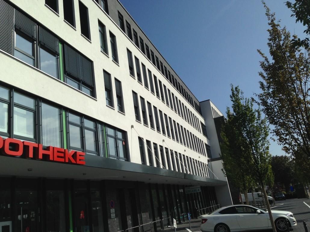 Weißes Gebäude mit „APOTHEKE“-Schild, schrägen Fenstern und einem draußen geparkten Auto an einem sonnigen Tag.