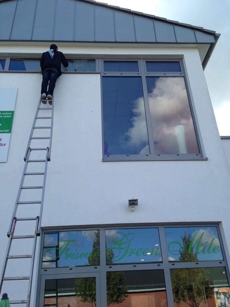 Mann auf einer Leiter putzt die Außenfenster eines weißen Gebäudes. Himmel und Wolken spiegeln sich im Glas.
