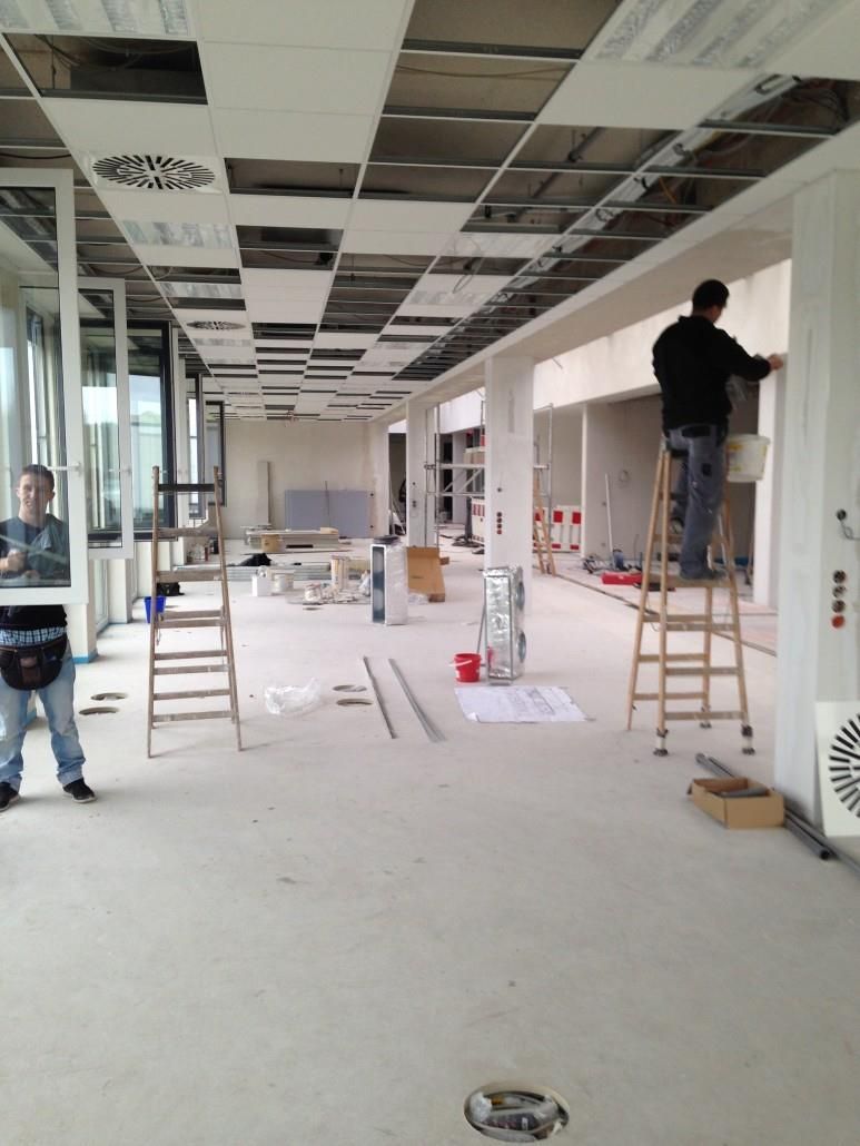 Innenansicht einer Baustelle. Zwei Männer arbeiten; einer malt, der andere steht. Weiße Wände, offene Decke.