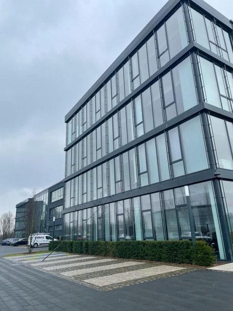 Modernes Bürogebäude mit Glaswänden und dunkelgrauer Fassade, bedecktem Himmel. Grünflächen im Erdgeschoss und Parkplätze vor dem Gebäude.