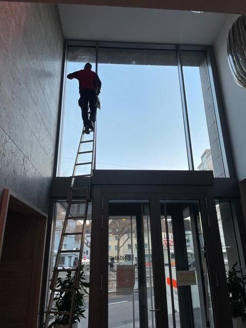 Person auf einer Leiter, die ein großes Fenster über einem Eingang reinigt. Gebäudeinneres, blauer Himmel.