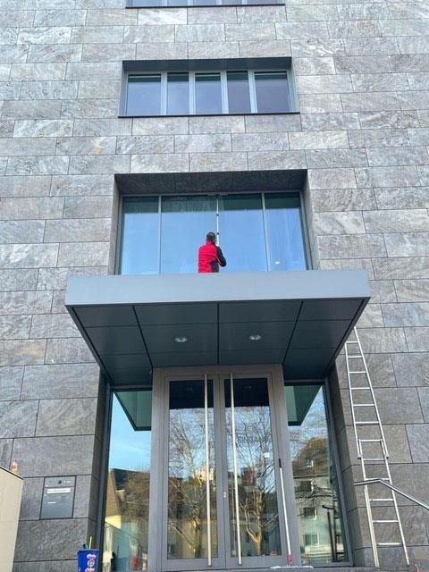 Person im roten Hemd putzt ein großes Fenster an einem Gebäude. In der Nähe steht eine Leiter.