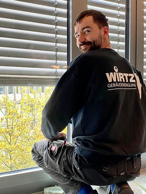 Mann in Arbeitskleidung kniet lächelnd am Fenster. Auf seinem Rücken befindet sich das Logo „WIRTZ Gebäudereinigung“.