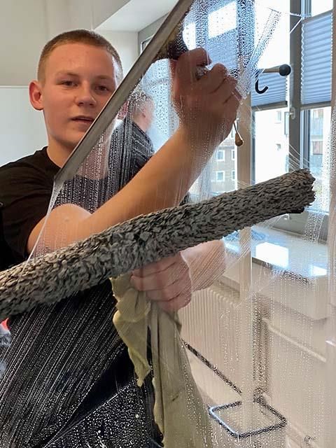 Junger Mann putzt drinnen ein Fenster mit Abzieher und Tuch.