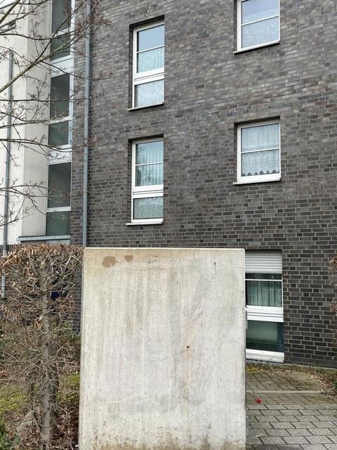 Graues Backsteingebäude mit weiß gerahmten Fenstern. Davor befindet sich ein großer Betonplatz.