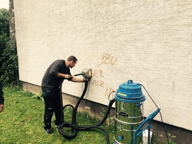 Mann entfernt Graffiti von einer Wand mit einem Sandstrahler und einem Staubsauger in einer grasbewachsenen Außenumgebung.