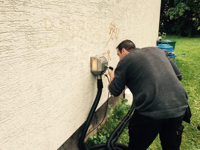 Mann arbeitet an Geräten, die mit Schläuchen und Grünpflanzen an einer beigen Außenwand befestigt sind.
