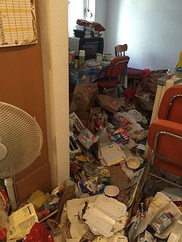 Ein überfüllter Raum voller Müll und Essensreste. Im Vordergrund steht ein orangefarbener Stuhl, daneben ein Ventilator.