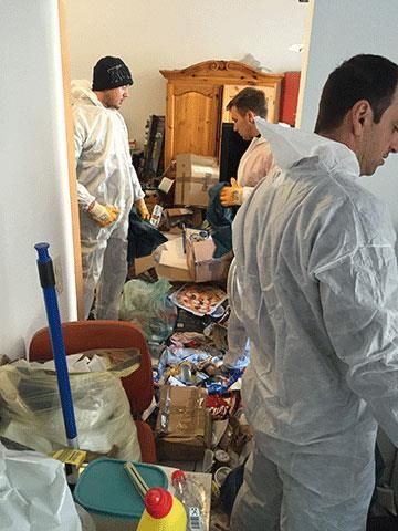 Drei Personen in weißen Schutzanzügen reinigen einen unordentlichen Raum.