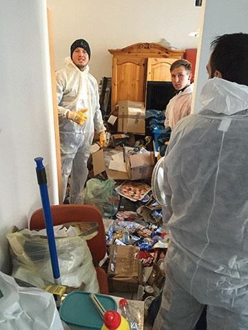 Drei Personen in Schutzanzügen in einem überfüllten Raum, Blick durch die Tür. Kartons und Müll liegen verstreut herum.