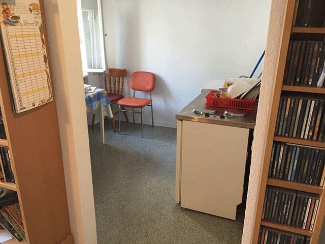 Blick durch die Tür in einen kleinen Küchen-/Essbereich: Stühle, Tisch, Schrank, Regale, Kalender.