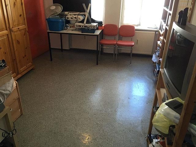 Zimmer mit grau gesprenkeltem Boden, Schreibtisch, Stühlen, Holzschrank und Regal mit Fernseher.