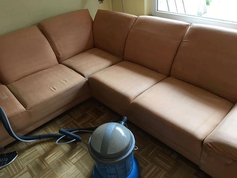 Eine Person saugt im Innenbereich ein helloranges Sofa neben einem Fenster ab.