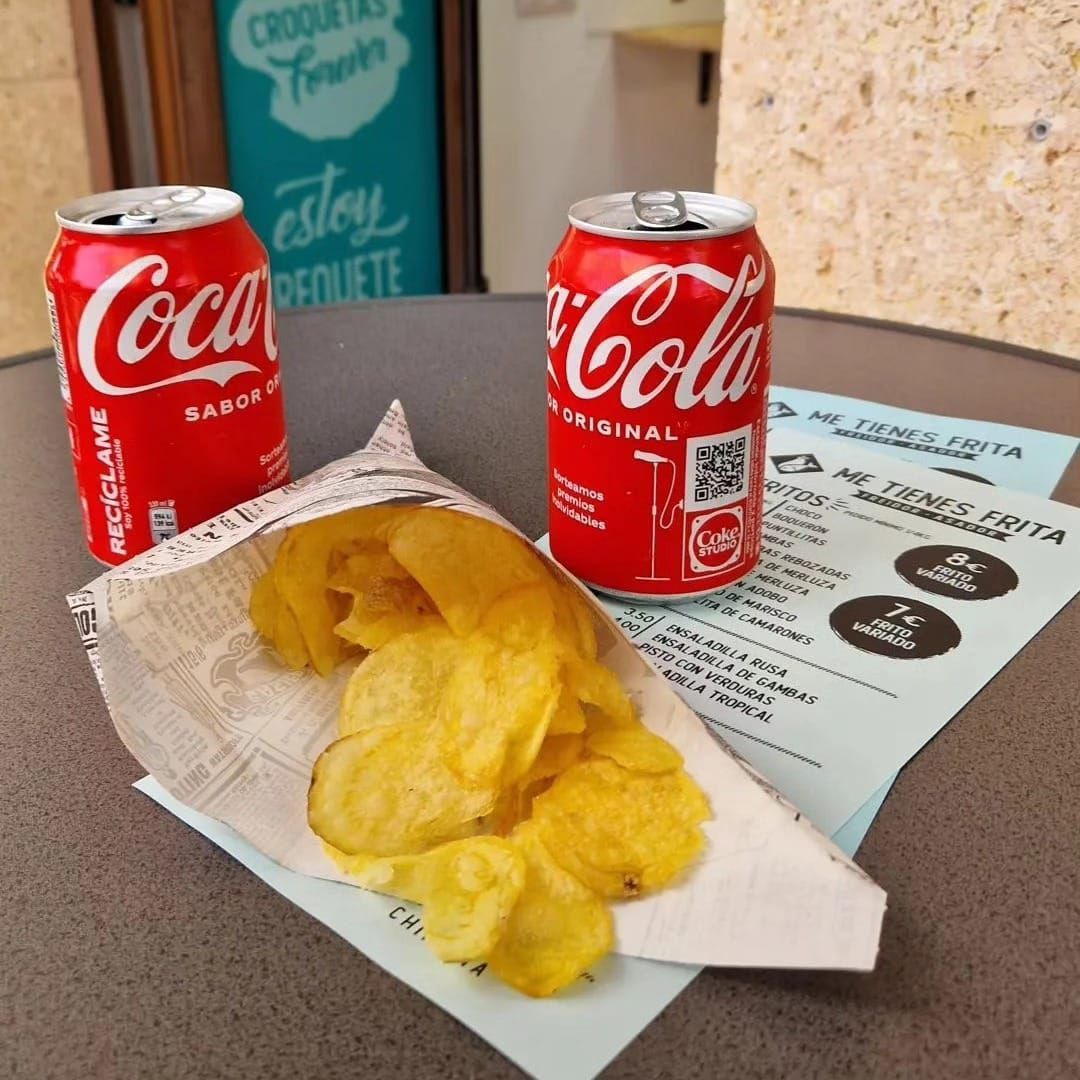 Dos latas de Coca-Cola, una bolsa de patatas fritas y un menú en una mesa al aire libre.