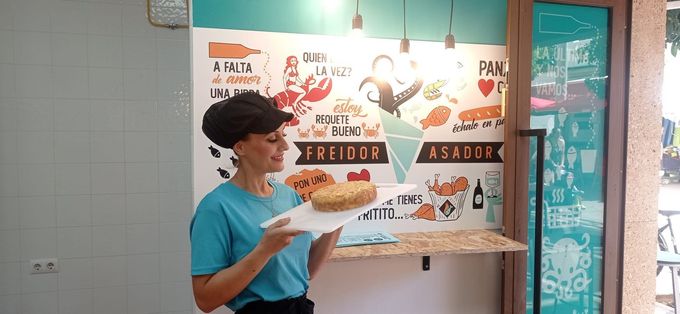Mujer con camisa y gorra verde azulado sosteniendo un pastel en una bandeja blanca, sonriendo. En una cafetería.