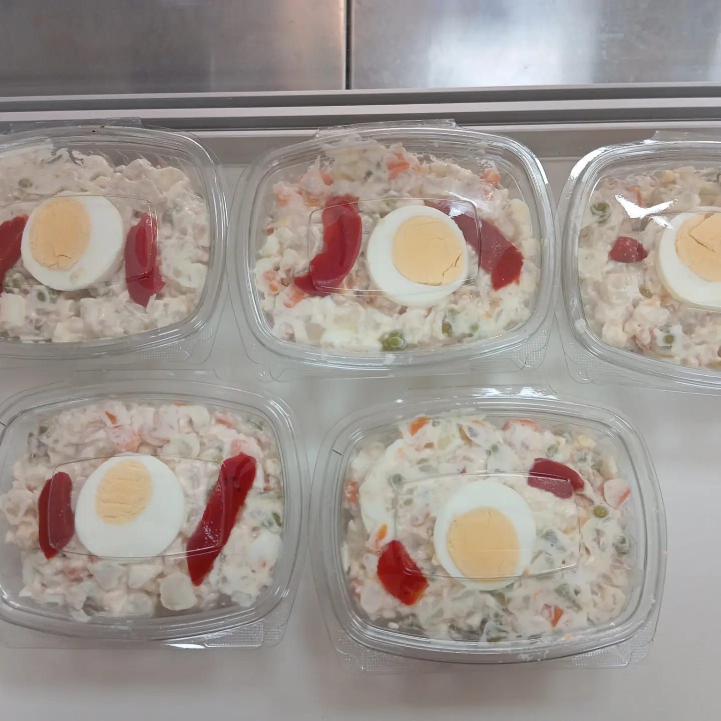 Cinco recipientes de plástico transparente con ensalada de patatas, cubiertos con huevo y pimiento rojo.