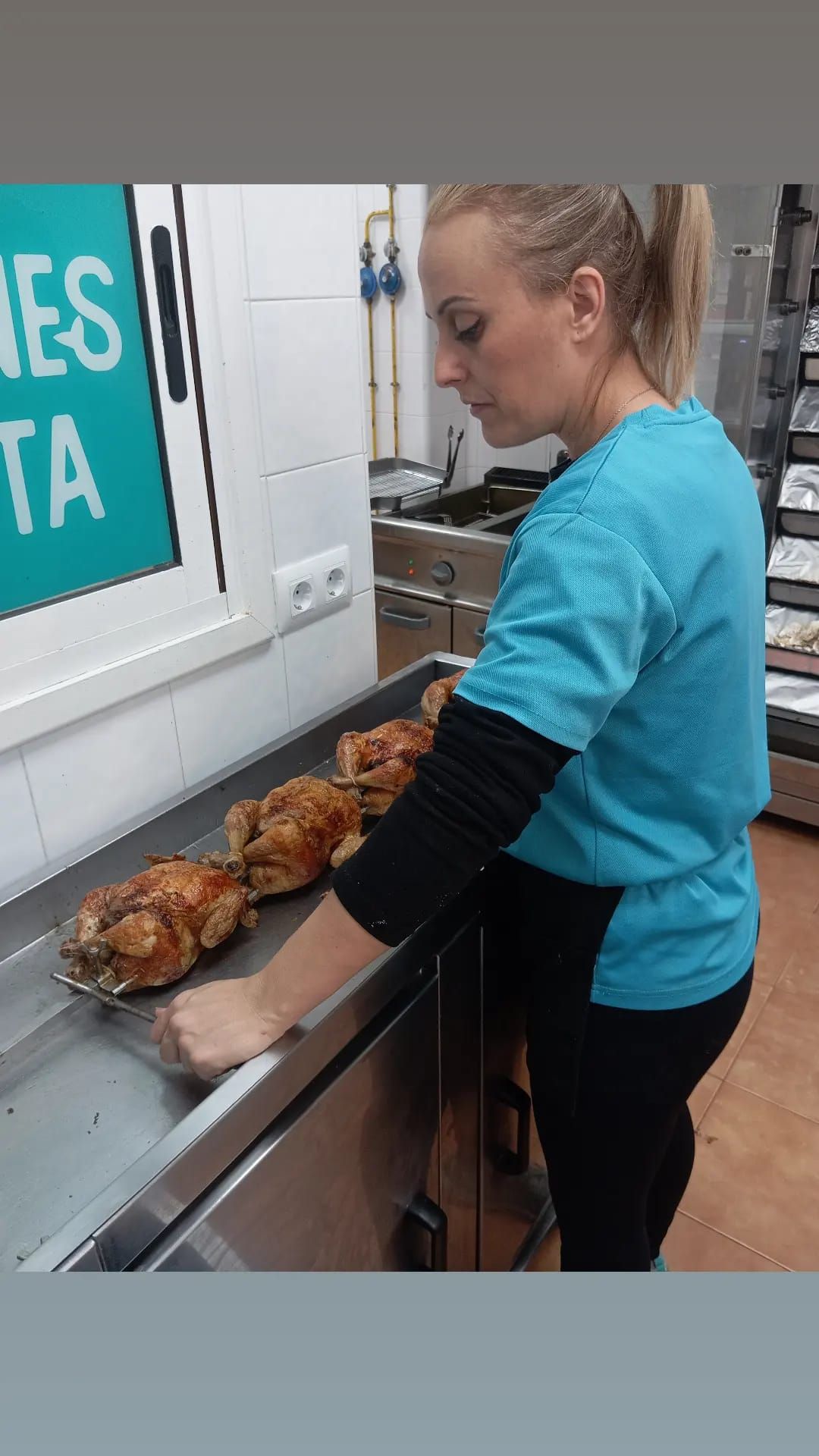 Mujer con camisa azul colocando pollos asados ​​en un mostrador de acero inoxidable en una cocina.