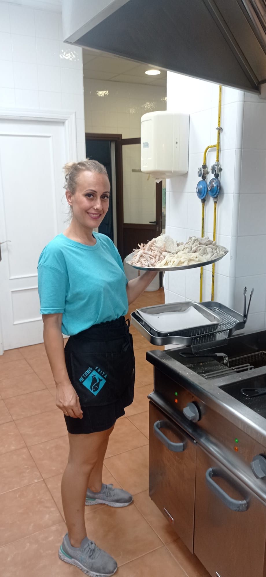 Mujer con una camisa azul y un delantal negro sosteniendo una pizza, de pie en una cocina.