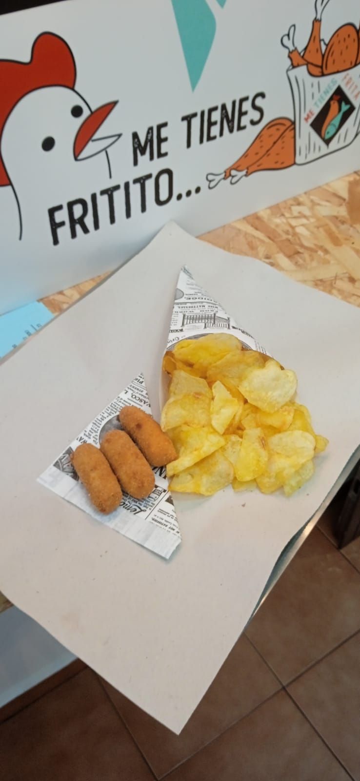 Comida frita en una bandeja forrada de papel: croquetas y patatas fritas, con un cartel con temática de pollo de fondo.