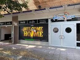 Außenansicht der Bar „Alpenrose“. Ein Willkommensbanner mit einem Bierglas ist zu sehen. Das Gebäude ist grau und weiß und hat Doppeltüren.