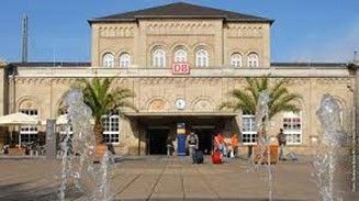 Vor einem Bahnhof in sonniger Umgebung, mit einem Brunnen im Vordergrund und Palmen am Eingang. Mehrere Personen befinden sich in der Nähe des Eingangs.