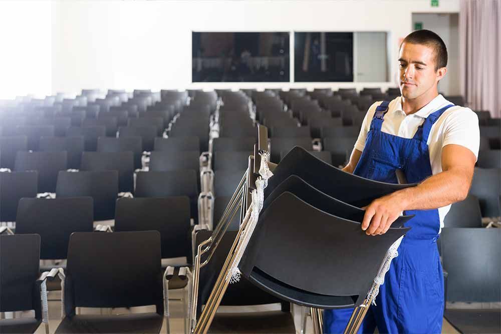 Ein Mann im blauen Overall trägt einen Stapel Stühle in einem Auditorium.