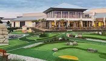 Minigolfplatz mit grünem Kunstrasen, einem Gebäude mit Restaurant und Landschaftselementen wie Felsen und Wasserspielen.