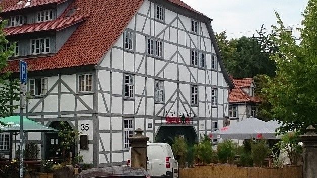 Ein Fachwerkhaus mit rotem Ziegeldach, weißen Wänden und grauen Balken. In der Nähe parkt ein Lieferwagen, und man sieht eine Terrasse mit Sonnenschirmen.