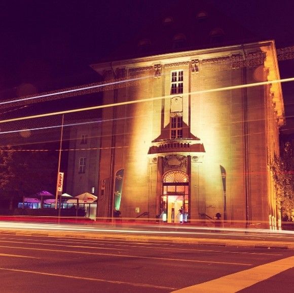 Ein nachts beleuchtetes Backsteingebäude mit langen Lichtstreifen vorbeifahrender Autos, einem beleuchteten Eingang und einem kleinen Sitzbereich im Freien auf der linken Seite.