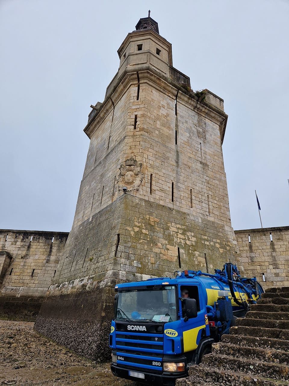 Camion bleu sur des marches au pied d'une tour en pierre, sur fond de ciel nuageux.